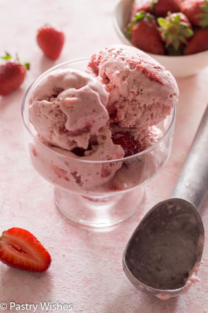 scoops of homemade strawberry ice cream in a bowl with strawberries and ice cream scoop on surface