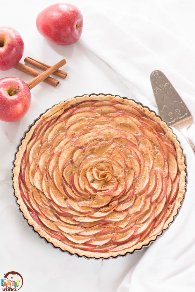 apple rose tart on a white countertop with two apples and cinnamon sticks and spatula